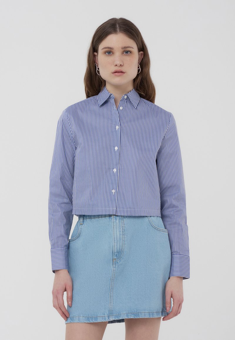 Camicia a maniche lunghe con bottoni, ritagliata, a righe verticali blu e bianche, con colletto, abbinata a una gonna di jeans blu chiaro.