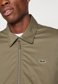 Tamsiai žalia zip-up striukė su krakmolu, kuriame kairėje pusėje yra maža siuvinėta krokodilo logotipas. Pagaminta iš lygaus, lengvo audinio.