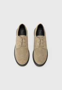 Chaussures en daim beige à lacets avec des détails brogue, surface texturée, orteil arrondi et semelle en caoutchouc noir contrastante. Logo de la marque visible à l'intérieur.