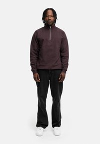 Dunkellila Zip-Up Sweatshirt, strukturierten Stoff, hoher Kragen, kombiniert mit schwarzen Jeans und weißen Sneakers, vor einem schlichten Hintergrund stehend.