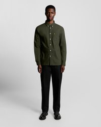 Camisa verde de manga larga con bolsillo en el pecho; presenta un pequeño logo. Combinada con pantalones de pierna ancha negros y zapatos negros.