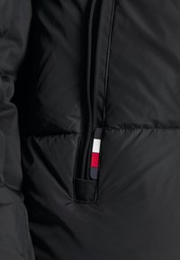 Veste matelassée noire avec une poche zippée équipée d'une tirette rectangulaire noire, blanche et rouge près de la couture.