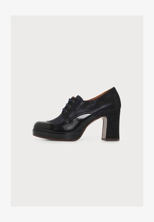 Scarpa nera da donna con tacco alto, chiusura frontale con lacci, texture miste di materiali lucidi e scamosciati, e tacco spesso.