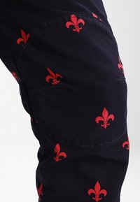 Černé látkové kalhoty s opakujícím se červeným vzorem fleur-de-lis, s viditelným šitím a přiléhavým střihem.