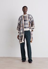 Poncho în carouri, în nuanțe de bej, albastru marin și maro, cu o croială lejeră, deschis în față și mâneci largi, asortat cu pantaloni drepți verzi.