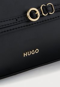 Mustast nahast kott, millel on kuldne logo ja metallosad, õmbluste detailid ning ümmargune tõmblukukinnituse aksent. Kompaktne ja sile tekstuur.