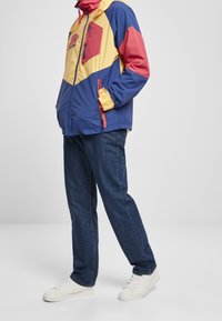 Kolorowa kurtka przeciwdeszczowa z panelami w kolorze niebieskim, żółtym i czerwonym, o sześciokątnym wzorze, z kieszeniami na zamek i elastycznymi mankietami. Dopełniona ciemnymi dżinsami i białymi trampkami.