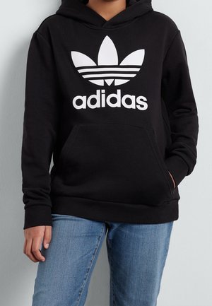 Osoba ubrana w czarną bluzę z kapturem Adidas z białym logo trefoil i napisem, w połączeniu z niebieskimi dżinsami, stojąca na tle jasnej ściany.