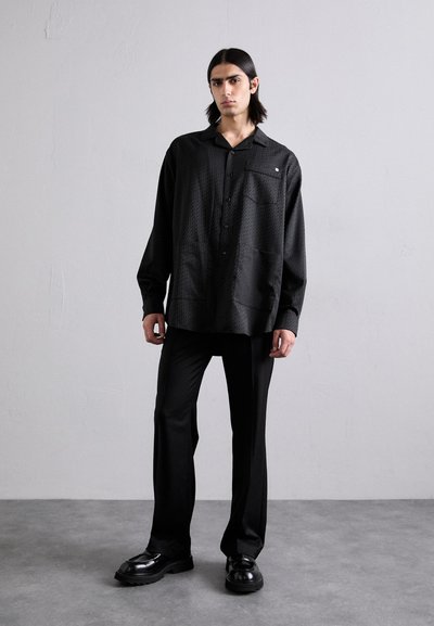 Schwarzes Hemd mit Knöpfen und strukturiertem Muster, kombiniert mit schwarzen Hosen und polierten schwarzen Schuhen, was einen monochromen Look schafft.