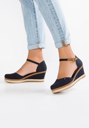 Fødder iført sorte kilehæle med ankelremme parret med lyseblå opprullede jeans på en hvid baggrund.