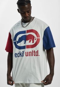 Šviesiai pilka trumpomis rankovėmis marškinėliai su raudonais ir mėlynais akcentais, su grafika, vaizduojančia raganosį, ir ryškiu tekstu "eckō unltd." priekyje.