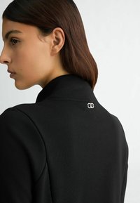 Crni visoki ovratnik od tkanine s teksturom i suptilnim detaljima šivanja, s malim srebrnim logotipom na stražnjoj strani.