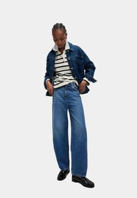 Blauwe denimjack over een gestreepte polo, gecombineerd met wijde blauwe jeans en zwarte instappers. Textuurstof en comfortabele pasvorm.
