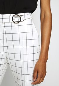 Pantalon blanc à carreaux avec un motif en grille noire, une large ceinture et un accent de boucle circulaire argentée. Tissu à texture lisse.