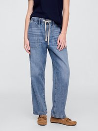 Persona, kas valkā gaiši zilos platajās biksēs ar elastīgu jostu un aukliņu, tumši zilu augšējo apģērbu un brūnas mokasīnu kurpes uz vienkārša fona.