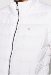 Biała kurtka puchowa z wysokim kołnierzem, suwakiem i poziomym pikowaniem. Posiada małą łatę z logo na piersi oraz błyszczący detal zamka.