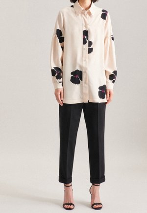 Persoon gekleed in een beige overhemd met grote zwarte bloemmotieven, zwarte omgeslagen broek en zwarte open-toe hakken, staand tegen een eenvoudige achtergrond.