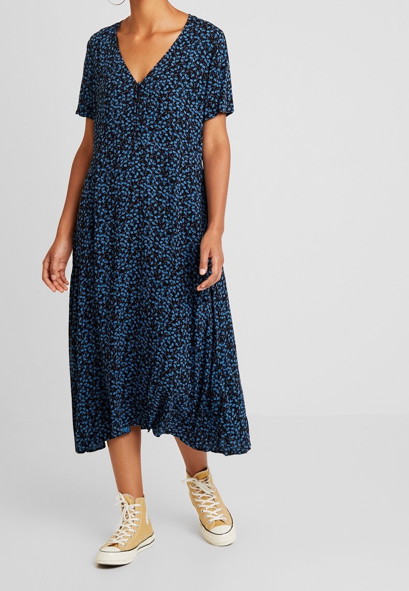 Femme vêtue d'une robe midi noire avec un petit imprimé floral bleu et des baskets montantes beiges, debout devant un fond uni.