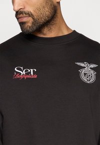 Fekete pulóver bordás nyakkal, fehér hímzett logóval és piros "Ser Benfiquista" felirattal a bal oldalon. Puha szövettextúra.