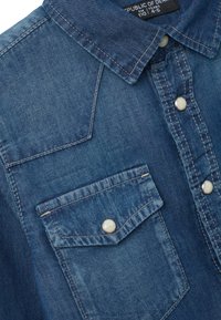 Primer plano del cuello de una camisa de mezclilla azul con un bolsillo cosido y botones de presión de perla blanca, etiquetada para niños de 4 a 5 años.