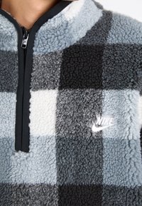 Fleece-pullover med halvdragkedja. Har ett mönster i svart, vitt och grått rutigt, mjuk textur och en liten vit Nike-logga.