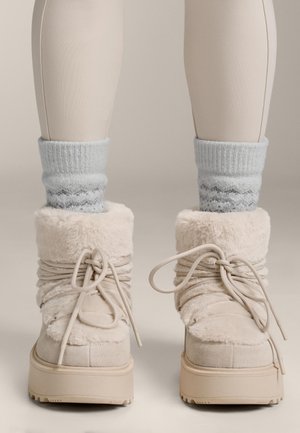Beige fur-lined boots with a plush texture, featuring a thick sole and soft, gathered shaft. Paired with light gray knitted socks.