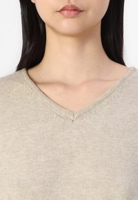 Młoda kobieta z długimi ciemnymi włosami, ubrana w beżowy sweter dzianinowy z dekoltem w kształcie litery V, pokazana od ramion w górę na białym tle.