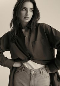 Ciemnobrązowa koszula zapinana na guziki z kieszenią na piersi, w połączeniu z jasnoszarymi spodniami o wysokim stanie i białym, prążkowanym topem pod spodem.