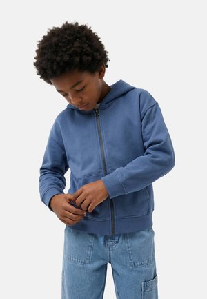 Garçon aux cheveux bouclés portant un sweat à capuche bleu à fermeture éclair et un pantalon en denim, concentré sur la fermeture du zip.