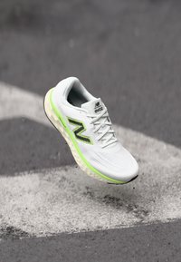 White athletic shoe with a textured upper, black and neon green accents, and cushioned sole, suspended above a gray surface.