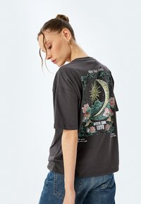 Donkergrijze katoenen T-shirt met een oversized snit. Heeft een kleurrijke grafische print van de zon, de maan en bloemen op de achterkant. Korte mouwen.