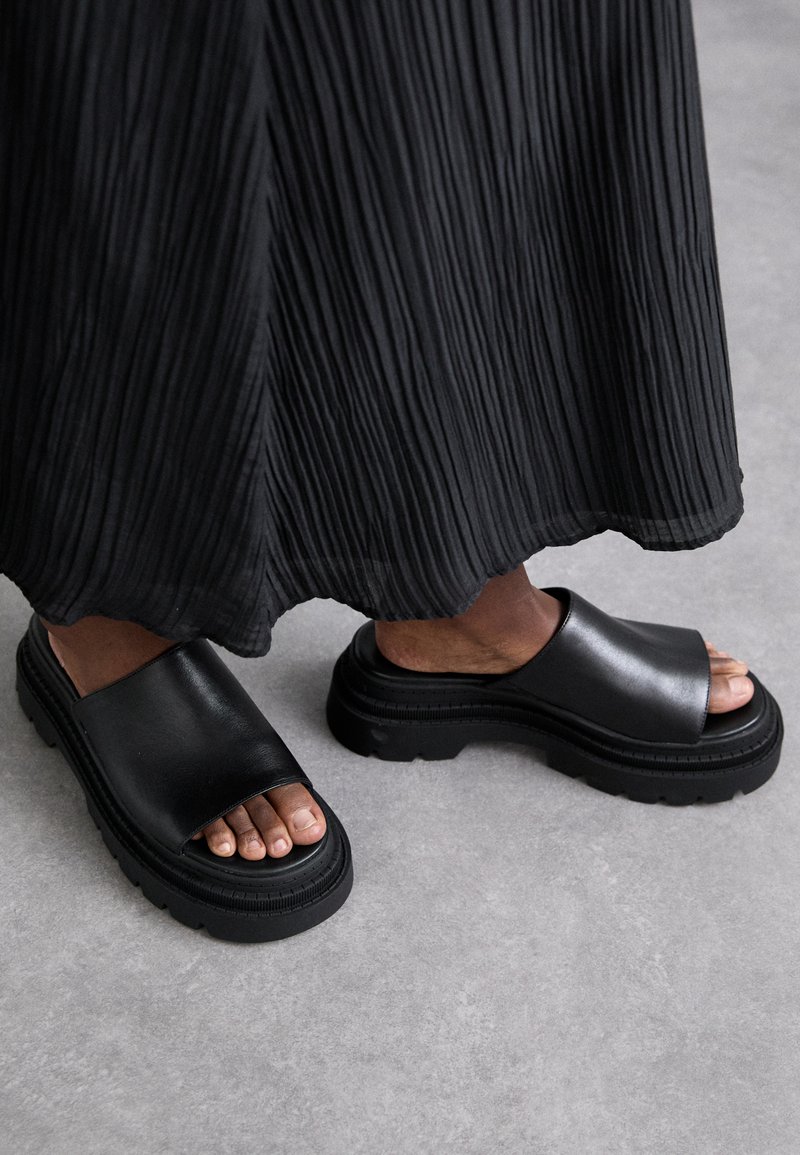 Sandalias deslizantes de cuero negro con diseño de punta abierta, suela gruesa y texturizada. Combinando con una falda negra plisada.