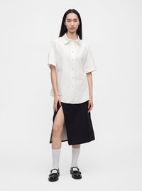 Femme portant une chemise blanche à manches courtes boutonnée, une jupe noire avec une fente haute à la cuisse, des chaussettes blanches et des mocassins noirs, debout devant un fond blanc.