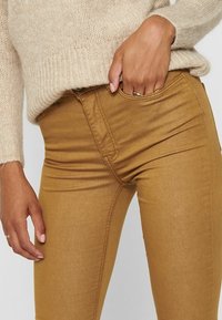 Bruine skinny jeans met een gladde textuur, voorzien van gedetailleerde stiksels, een middelhoge taille en zijzakken. Gecombineerd met een lichtgekleurde gebreide trui.