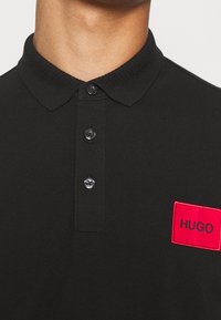 Homme portant un polo noir avec trois boutons et un patch rectangulaire rouge portant la mention "HUGO" sur la poitrine.