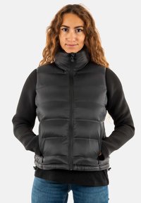 Gilet matelassé noir avec col haut, fermeture éclair à l'avant et ourlet élastique. Le gilet présente un design matelassé et des poches latérales. Porté par-dessus une chemise noire à manches longues.