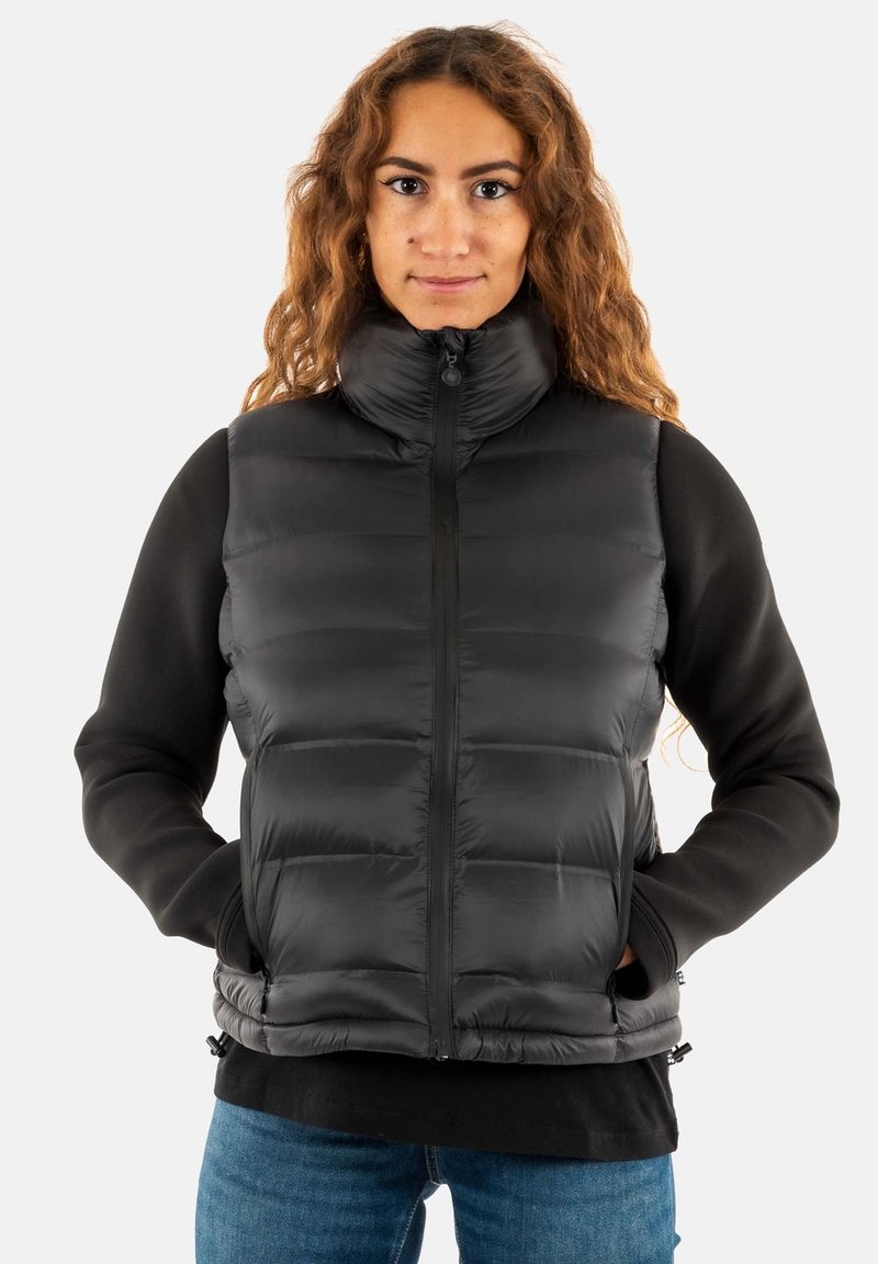 Gilet matelassé noir avec col haut, fermeture éclair à l'avant et ourlet élastique. Le gilet présente un design matelassé et des poches latérales. Porté par-dessus une chemise noire à manches longues.