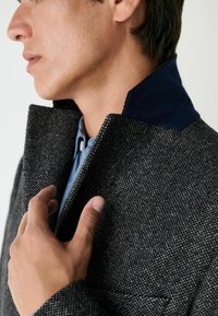 Gyapjú keverék kabát szürke houndstooth mintával, tengerészkék velúr gallérral és textúrált anyaggal, magas gallérral és cipzáros részletekkel.