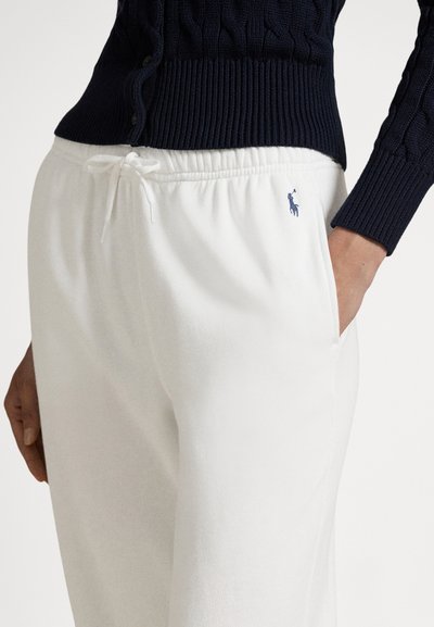 Fehér tréningnadrág gumis derékrésszel és húzózsinórral. Bal csípőjén egy kis kék logó található. Hozzá egy tengerészkék bordázott pulóver párosítva.