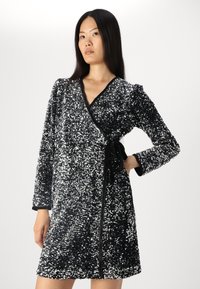 Μαύρο και ασημί φόρεμα με παγιέτες, με σχέδιο τύπου περιτυλίγματος, μακριά μανίκια και ζωνάκι. Η υφή του αστράφτει, προσφέροντας ένα ανακλαστικό εφέ.