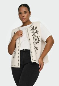 Femme portant un t-shirt blanc et un jean noir avec un gilet sans manches crème orné de broderies florales noires, posant avec une main sur la poitrine.