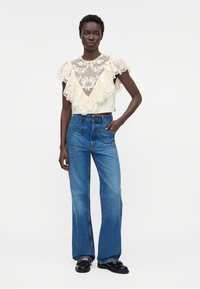 Femme portant un crop top en dentelle crème à volants, un jean bleu taille haute avec poches avant et des mocassins noirs, debout avec une main dans la poche.