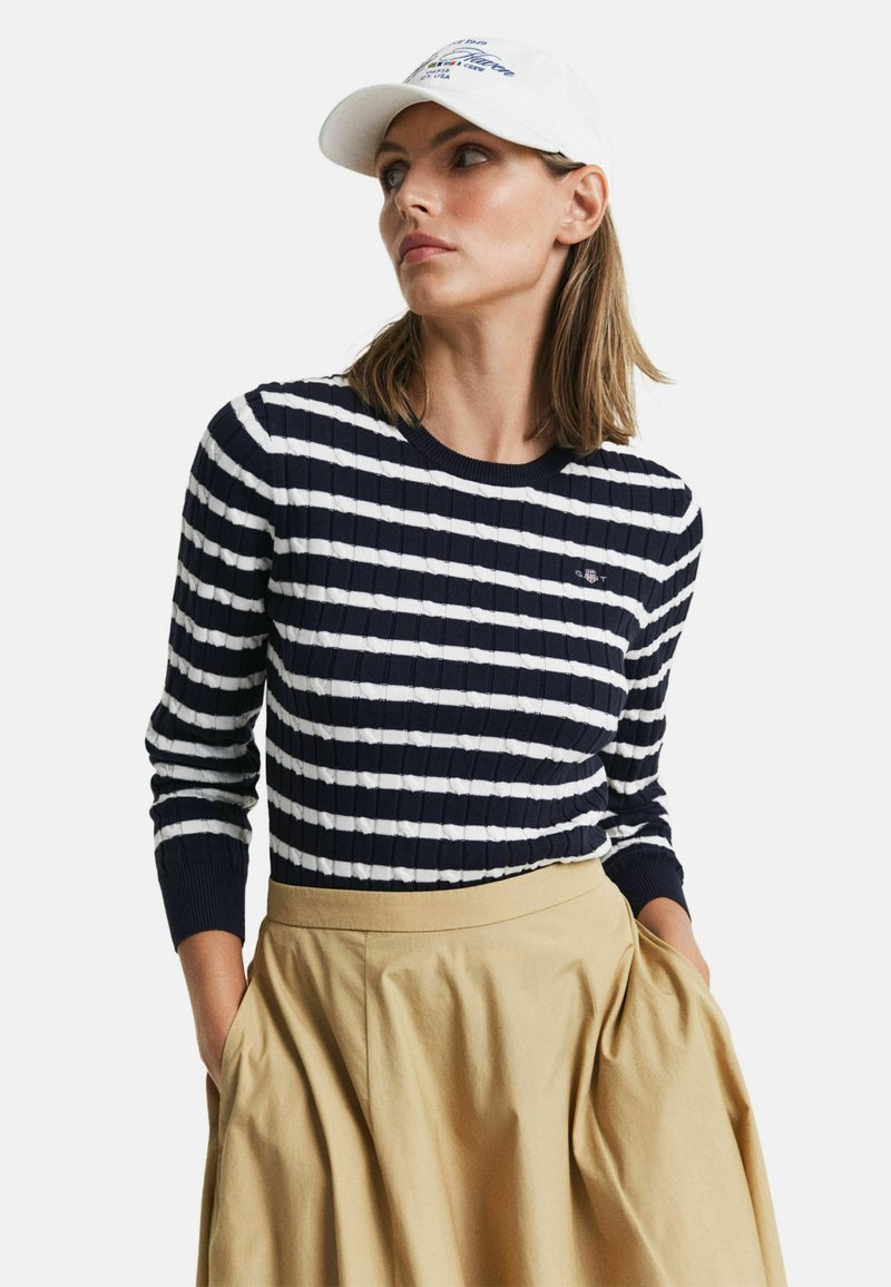 Blau-weißer gestreifter Ripp-Pullover mit langen Ärmeln, kombiniert mit einem beigen, plissierten Rock. Weiße Kappe mit Logodetails.