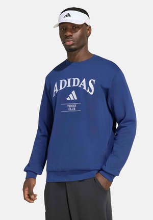 Mann trägt einen blauen Adidas Tennis Club Sweatshirt und schwarze Hose, trägt auch ein weißes Adidas Visier, steht mit einer Hand in der Tasche.