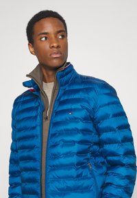 Blauwe pufferjas met een gestikte design en hoge kraag, voorzien van rode en witte accentstrepen en een ritsluiting.