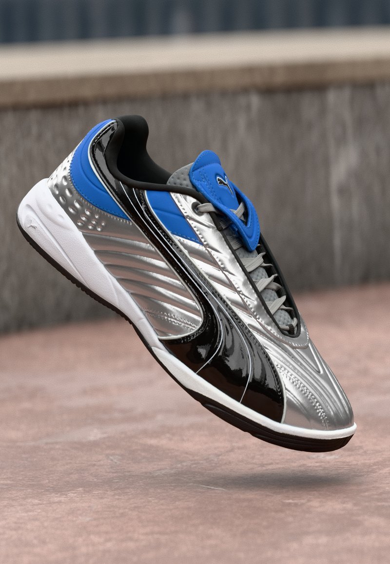 Sneaker sportivo argento e blu con dettagli neri e suola bianca, sospeso sopra una superficie di cemento rosa con uno sfondo sfocato di un muro grigio.