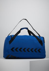Bolso de deporte azul con un patrón de flechas negras, hecho de un tejido duradero, que cuenta con dos asas negras y una correa de hombro desmontable.