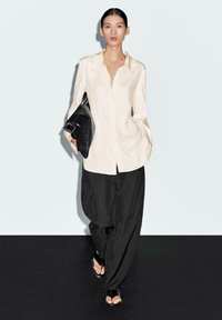 Camicia a maniche lunghe crema con chiusura a bottoni, abbinata a pantaloni neri larghi. Borsa nera con dettaglio a fibbia. Il modello indossa sandali neri.