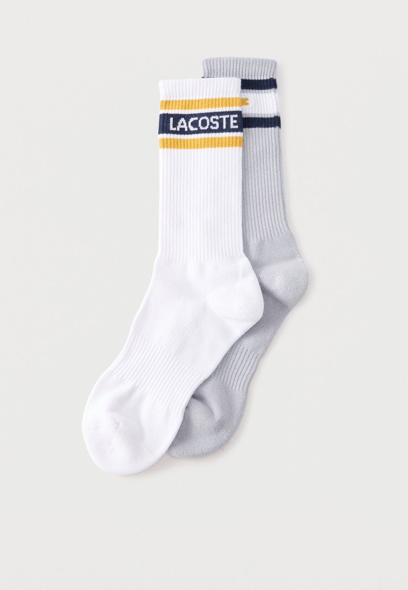 Paquet de deux paires de chaussettes côtelées, une blanche avec des rayures jaunes et bleu marine et le texte « LACOSTE », une gris clair avec des rayures bleu marine au bord-côte.