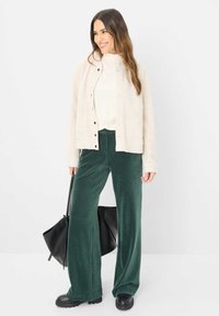 Lys beige button-up jakke over en tætsiddende hvid top, parret med brede grønne fløjlstrættebukser og sorte kraftige støvler med tyk sål. Sort håndtaske inkluderet.